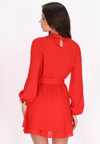 Rode jurk met lange mouwen, textuurstof en een ceintuur in de taille. Heeft een keyhole rugontwerp en een gelaagde zoom.