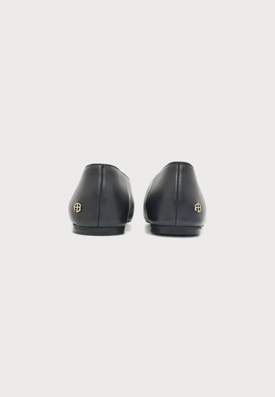 Paire de chaussures plates en cuir noir vues de dos, chacune avec un petit emblème rond doré près du talon.