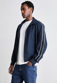 Jachetă navy cu fermoar, cu accente albe, manșoane ribbate și un design elegant. Purtați peste un tricou alb și asociată cu blugi albaștri.