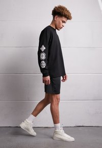 Black long-sleeve shirt with white graphic designs on the sleeves, paired with dark shorts and white sneakers, standing on a gray floor.