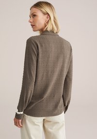 Une femme aux cheveux blonds courts porte un haut texturé marron, de dos, associé à un pantalon clair, le tout sur un fond neutre.