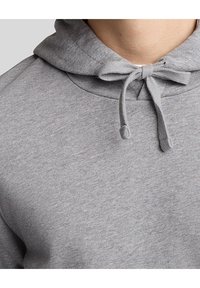 Grå huvtröja med en mjuk, texturerad tyg, snörningsdetalj och avslappnad passform. Närbild av halsringningen och huvan.