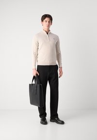 Suéter beige con cremallera, pantalones negros ajustados, sosteniendo un bolso de cuero negro, con calzado negro. Líneas simples y limpias en colores neutros.