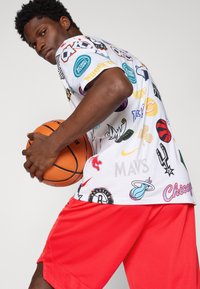 Bílý tričko se spoustou barevných sportovních log; v kombinaci s jasně červenými šortkami; osoba držící basketbal, ukazující svalnatou ruku.