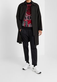 Schwarzer Mantel über einem roten Grafiksweater mit urbanen Designs, kombiniert mit dunkler Hose und weißen Sneakers mit schwarzen Akzenten.