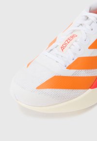 Biela športová obuv so svetlooranžovými diagonálnymi pruhmi, štruktúrovaným zvrškom a svetlou podrážkou s logom na jazyku.