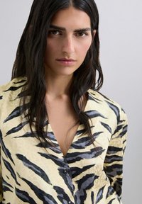 Camisa amarilla con rayas de cebra negras, que presenta un escote en V profundo y cierre de botones; la tela parece suave con una textura ligera.