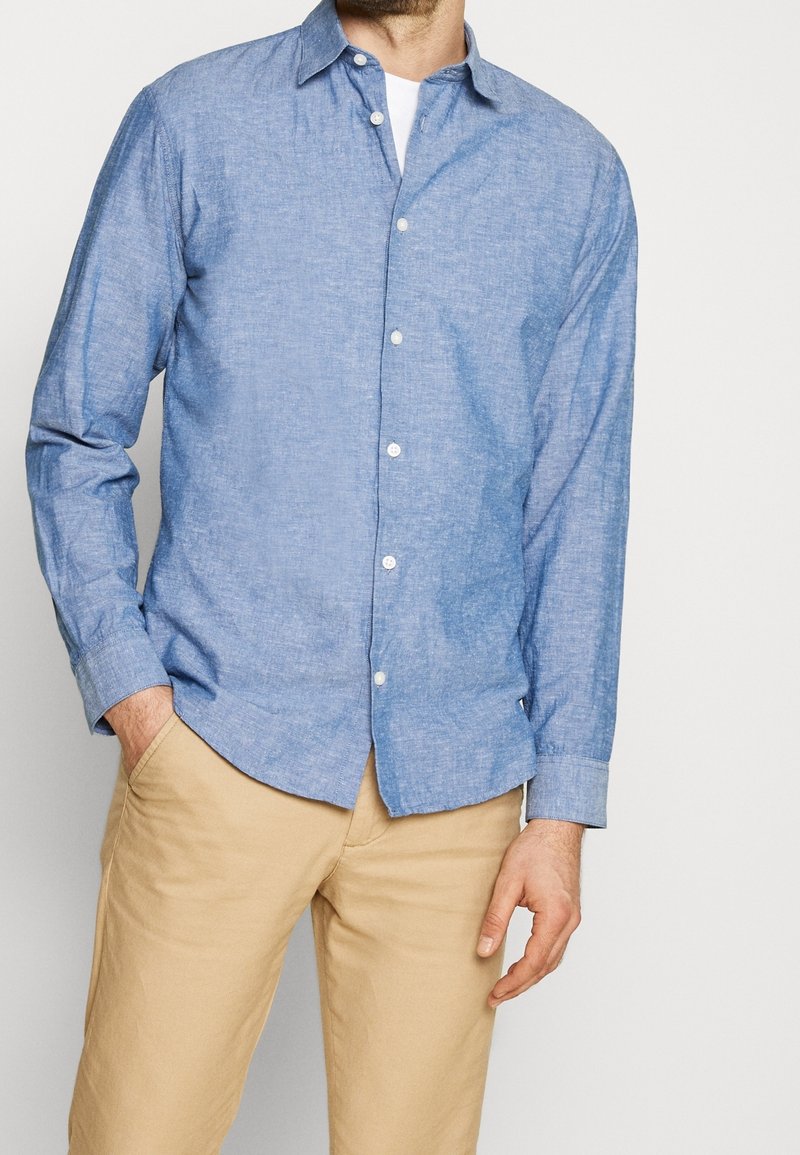 Homme portant une chemise bleu clair boutonnée sur un sous-vêtement blanc avec un pantalon beige, une main dans la poche, debout devant un fond uni.