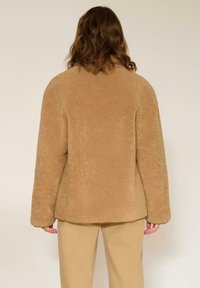 Veste en polaire beige et duveteuse, à coupe décontractée et manches longues. Elle présente un ourlet arrondi et une texture douce pour la chaleur.