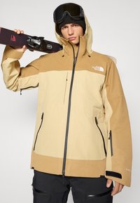 Beize ja pruun suusajope, millel on lukuga esiosa ja kaks külge taskut, valmistatud tekstureeritud kangast. The North Face logo on rinnal.