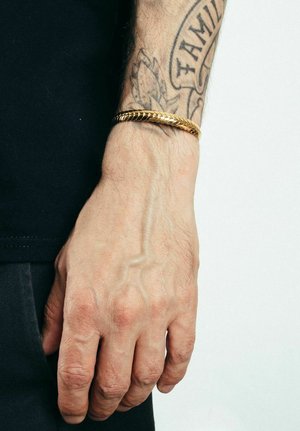 Pulsera trenzada de oro en una muñeca morena, con un diseño texturizado. La mano es parcialmente visible, con un tatuaje en el antebrazo.