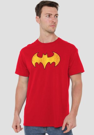 Man wearing red t-shirt with a large yellow bat symbol on the chest, standing against a plain light background.
