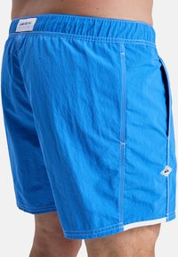 Blue swim trunks made of lightweight fabric with a textured finish. Features white stitching, side pocket, and logo patch on the hem.