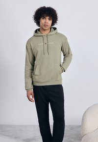 Mladý muž s kučeravými vlasmi, obliekajúci olivovo zelenú mikinu Tommy Hilfiger a čierne nohavice, stojí s jednou rukou vo vrecku.