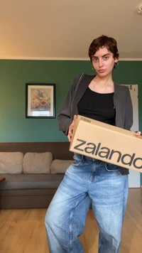 Kartiņu kaste ar uzrakstu "zalando", ko tur rokās cilvēks pelēkā jakā un melnā augšpusē, ar gaiši ziliem platām biksēm. Fons - dīvāns.