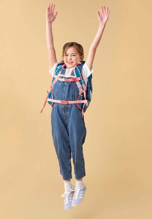 Girl jumping with arms raised, wearing striped overalls, white shirt, sneakers, and a blue backpack with pink straps, beige background.
