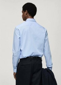 Cămașă de culoare albastru deschis, cu dungi, cu mâneci lungi, cu manșete buton, și cu un guler clasic, purtată cu pantaloni închiși la culoare și un pulover bleumarin așezat pe braț.