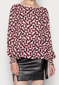 Gemusterte Bluse mit burgunderrotem, schwarzem und cremefarbenem geometrischem Design. Mit langen Ärmeln und gerafften Bündchen sowie einem weiten Ausschnitt. Kombiniert mit einem schwarzen Lederrock.