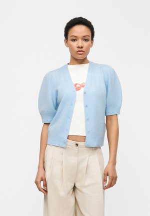 Kvinde iført en lyseblå cardigan med korte ærmer og knapper over en hvid crop top med rødt motiv og beige, højtaljede, plisserede bukser.