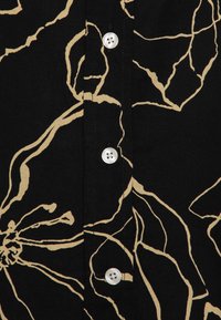 Svart skjorta med beige blommönster, kort knappslå och fyra vita knappar. Har ett texturerat tyg med synliga sömmar.