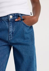 Blue denim jeans with a vertical stripe pattern, featuring a silver button closure and side pockets. Fabric has a smooth texture.