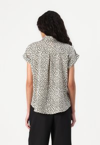 Blusa a maniche corte di colore crema con macchie astratte nere, dotata di colletto e dettaglio plissettato sul retro, realizzata in tessuto leggero.
