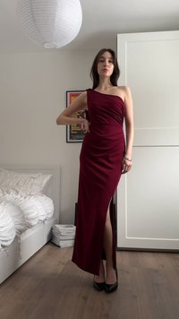 Nő bordó, egyrészes vállpántos ruhában, combvágással, fehér ágyneművel, fa padlóval és keretezett faliképpel berendezett hálószobában áll.