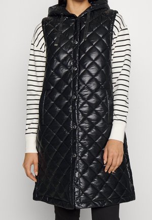 Femme portant une veste matelassée noire sans manches avec capuche sur une chemise à manches longues blanche et noire à rayures.