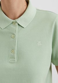 Lichtgroene polo shirt van een gestructureerde stof, met een drie knopen placket en een geborduurd "MOP" logo op de linkerborst.