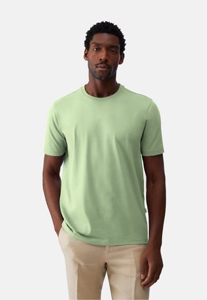 Uomo con maglietta verde chiaro a maniche corte e pantaloni beige, in piedi con una mano in tasca su uno sfondo semplice.