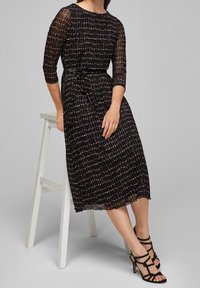 Falda negra midi con mangas three-quarter, con un patrón geométrico en beige y rosa, ceñida a la cintura con un lazo. Tacones negros con tiras.