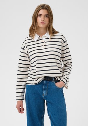 Jeune femme aux cheveux châtain clair portant une chemise blanche à manches longues avec des rayures horizontales noires et un jean bleu sur un fond uni.