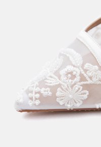 Κοντινό πλάνο λευκού πλεκτού παπουτσιού με κεντημένα λουλουδένια σχέδια και μικρές λεπτομέρειες με χάντρες στη μυτερή μύτη.