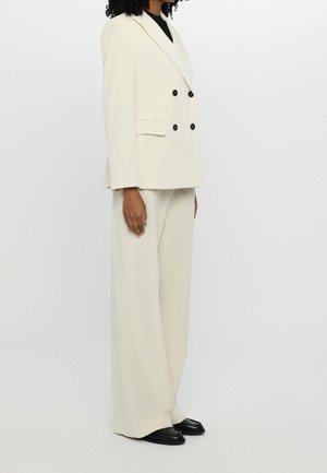 Woman wearing a cream double-breasted blazer and matching wide-leg pants with black shoes on a plain background.