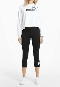 Kobieta w białej, krótkiej bluzie Puma, czarnych, krótkich legginsach Puma i białych sneakersach, stojąca na tle białym.