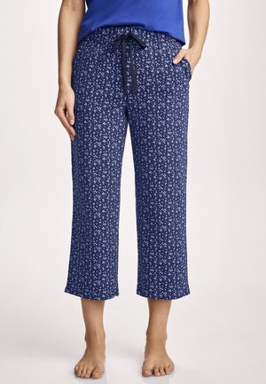 Kobieta w granatowych krótkich spodniach z drobnym, jasnoniebieskim kwiatowym wzorem i granatowym sznurkiem, stojąca boso na jasnym tle.