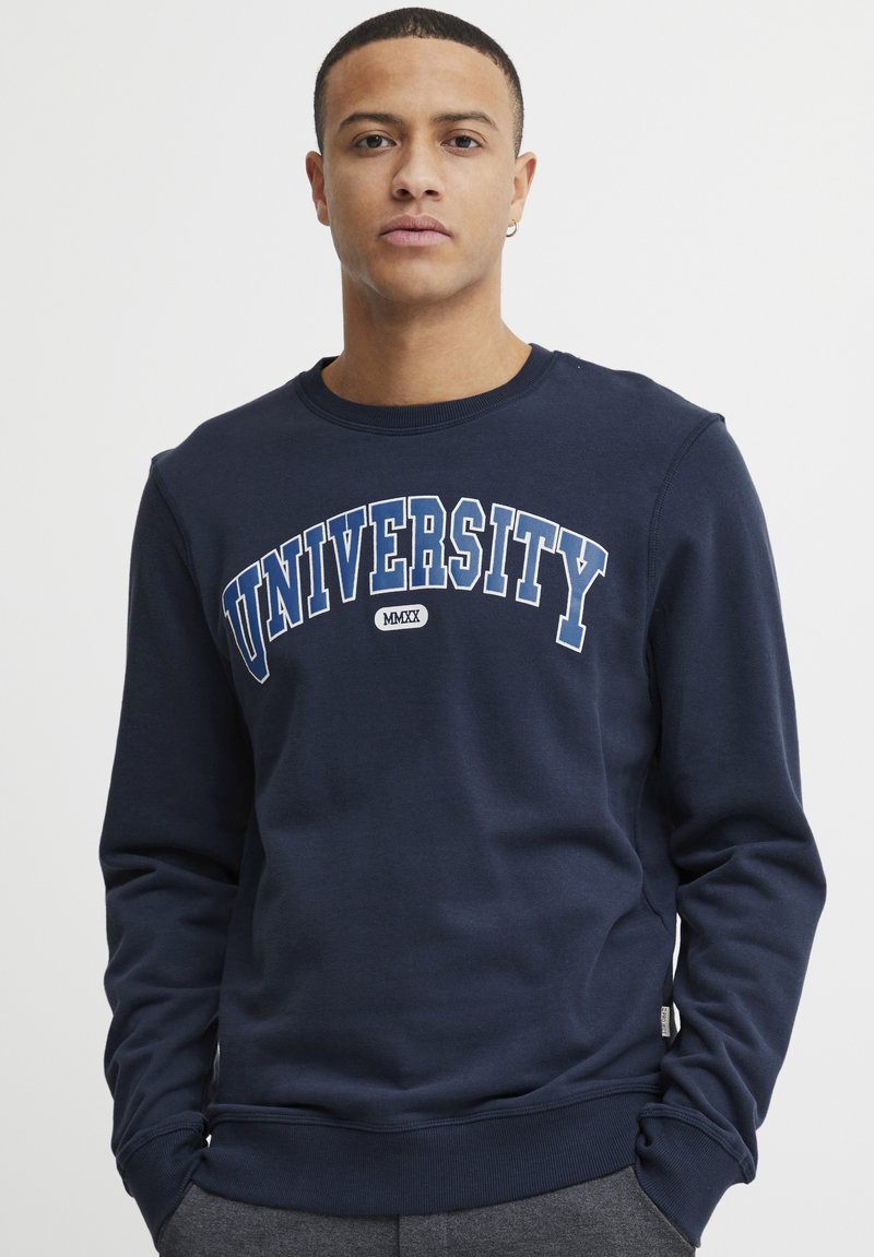 Marinblå sweatshirt med "UNIVERSITY" i vit och blå text. Ribbad crew-neck och ärmslut, mjuk textur, avslappnad passform.