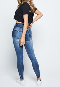 Hubené modré džíny s vybledlou texturou, odřenými akcenty na kapsách a surovými lemy, spárované s černým zkráceným tričkem a bílými botami.