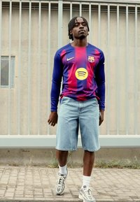 Dugih rukava plava i crvena prugasta majica s logom Barcelone i Spotify grafikom, uparen s svijetlim traper kratkim hlačama i sivim tenisicama.