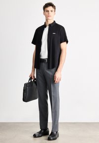 Μαύρο πουκάμισο με κοντά μανίκια και λογότυπο, λευκό μπλουζάκι, γκρι παντελόνι, μαύρη ζώνη, μαύρα δερμάτινα παπούτσια, κρατώντας μια μαύρη δερμάτινη τσάντα.