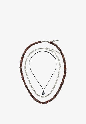 Trois colliers superposés : extérieur avec des perles en bois brun foncé, milieu avec des maillons de chaîne en argent, intérieur avec un cordon noir et un pendentif en pierre noire.