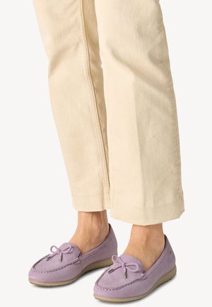 Persoon die beige broek draagt en lavendelkleurige suède mocassins met strikaccenten en beige rubberzolen, staand op een lichte achtergrond.