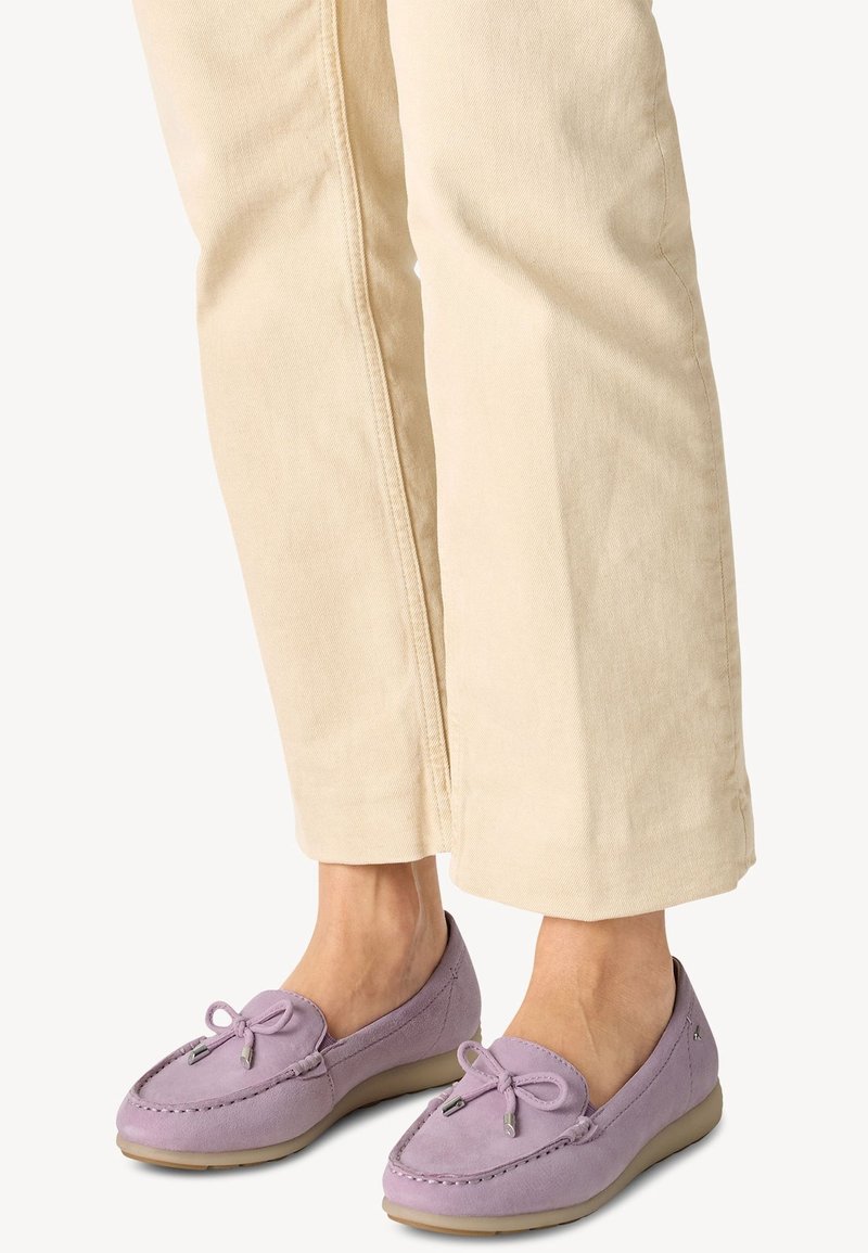 Personne portant un pantalon beige et des mocassins en daim lavande avec des accents de nœud et des semelles en caoutchouc beige, debout sur un fond clair.