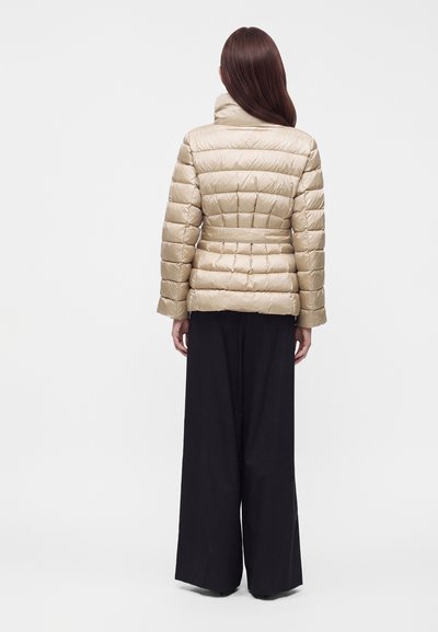 Femme aux longs cheveux bruns portant une veste matelassée beige et un pantalon large noir, debout de dos sur un fond uni.