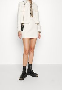 Biała kurtka z teksturowanej tkaniny z kieszeniami, kwiatowa bluzka, biała spódnica i czarne botki za kostkę z złotymi akcentami; elegancki i nowoczesny wygląd.