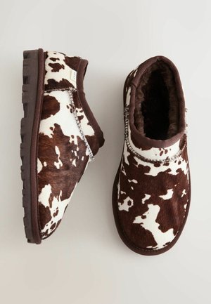 Brown and white cow print slip-on boots with a soft fur lining, rubber sole, and contrasting stitching detail on the seams.