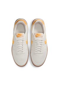 Valkoiset Nike-tennarit keltaisilla yksityiskohdilla, litteillä nauhoilla ja kumipohjalla. Sileä pinta, pyöristetty varvas ja näkyvä brändäys kielellä ja sivuilla.