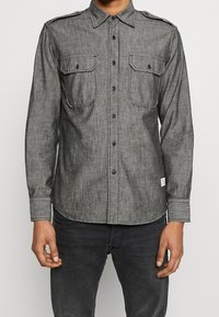 Uomo che indossa una camicia grigia con bottoni, dotata di due tasche sul petto e spalline, abbinata a jeans grigi scuri, in piedi con le braccia rilassate.