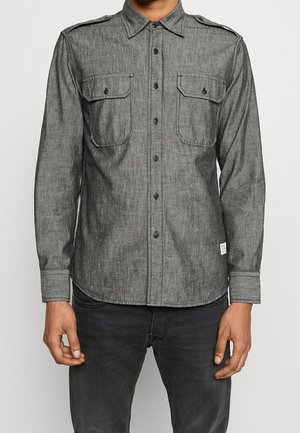Homme portant une chemise grise à boutons avec deux poches poitrine et des épaulettes, associée à un jean gris foncé, se tenant avec les bras détendus.