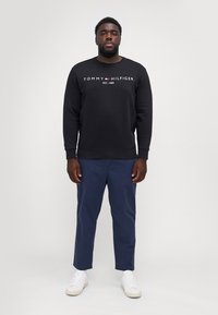 Black sweatshirt with "TOMMY HILFIGER EST. 1985" logo, paired with navy chinos and white sneakers. Smooth fabric, casual fit.
